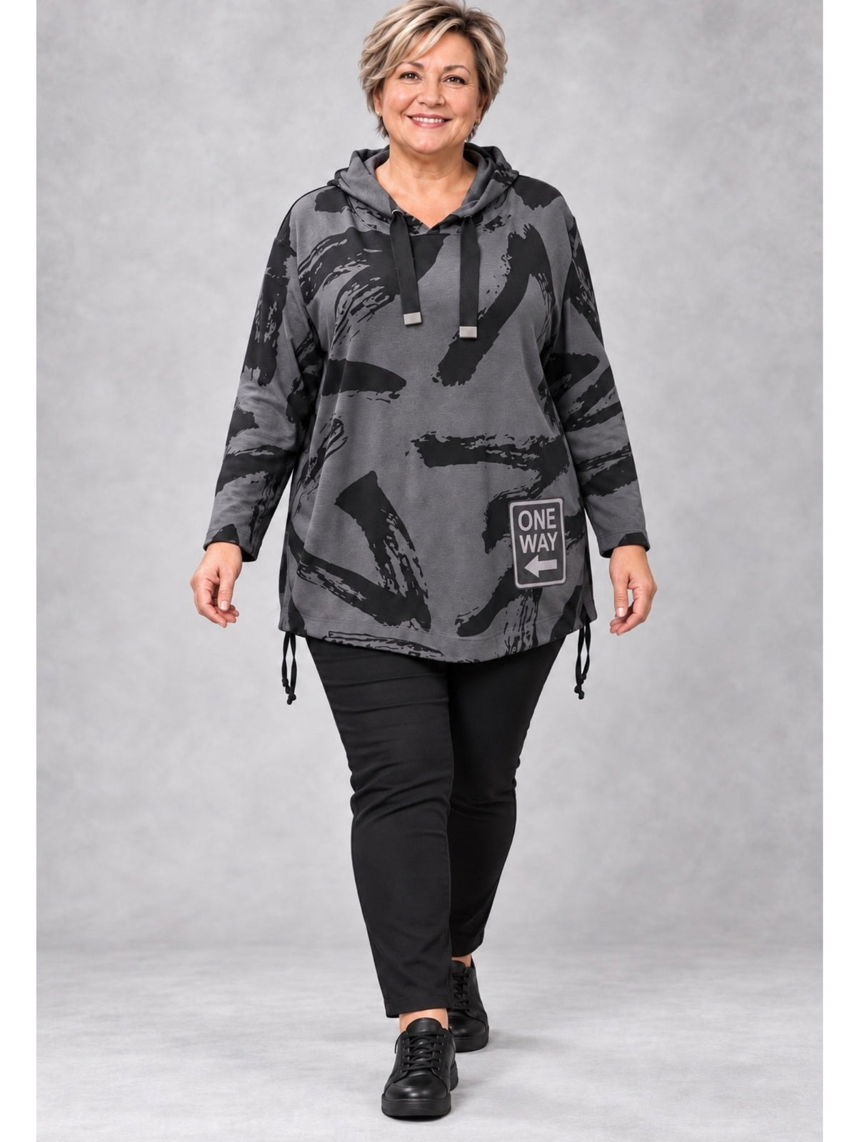 Graues Kapuzenshirt mit schwarzen Pinselstrichen, getragen mit schwarzer Hose und flachen Schuhen